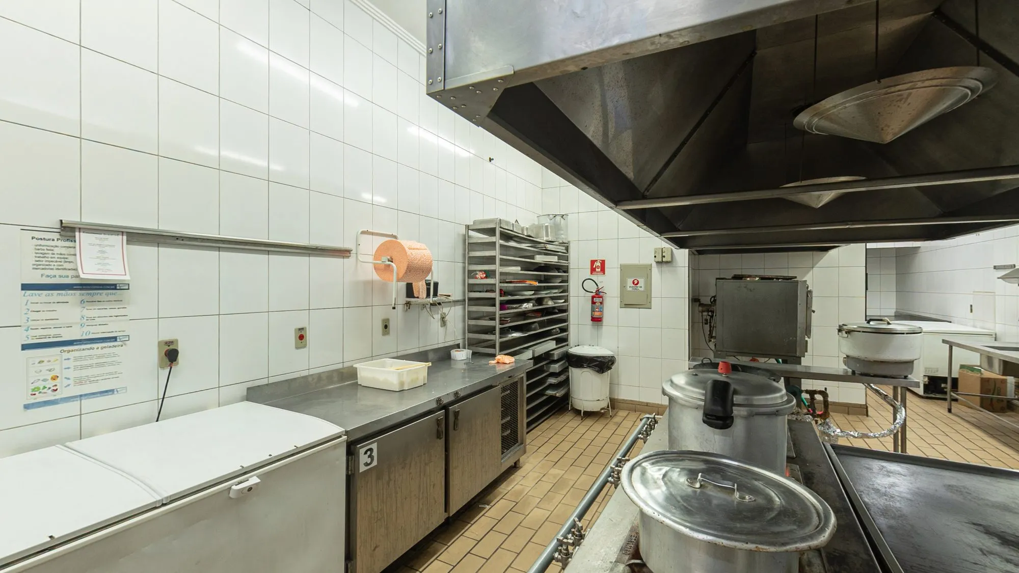 Cozinha profissional da Limelight, com paredes de azulejo branco, bancadas e fogão de aço inox, coifa, panelas grandes, freezers e um carrinho de assadeiras