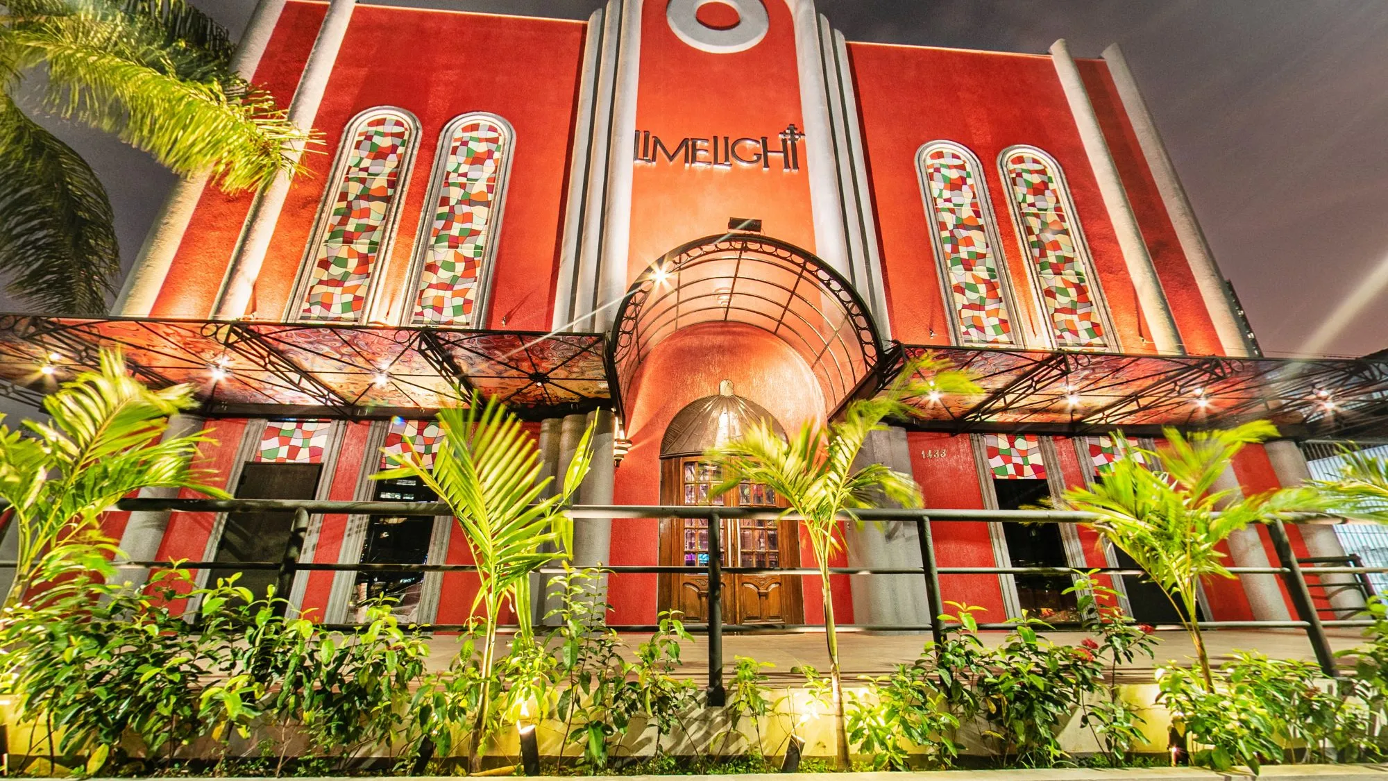 Fachada da Limelight à noite, com paredes vermelhas, janelas altas em arco com vitrais, o letreiro da casa, a marquise da entrada e palmeiras e canteiros iluminados na frente