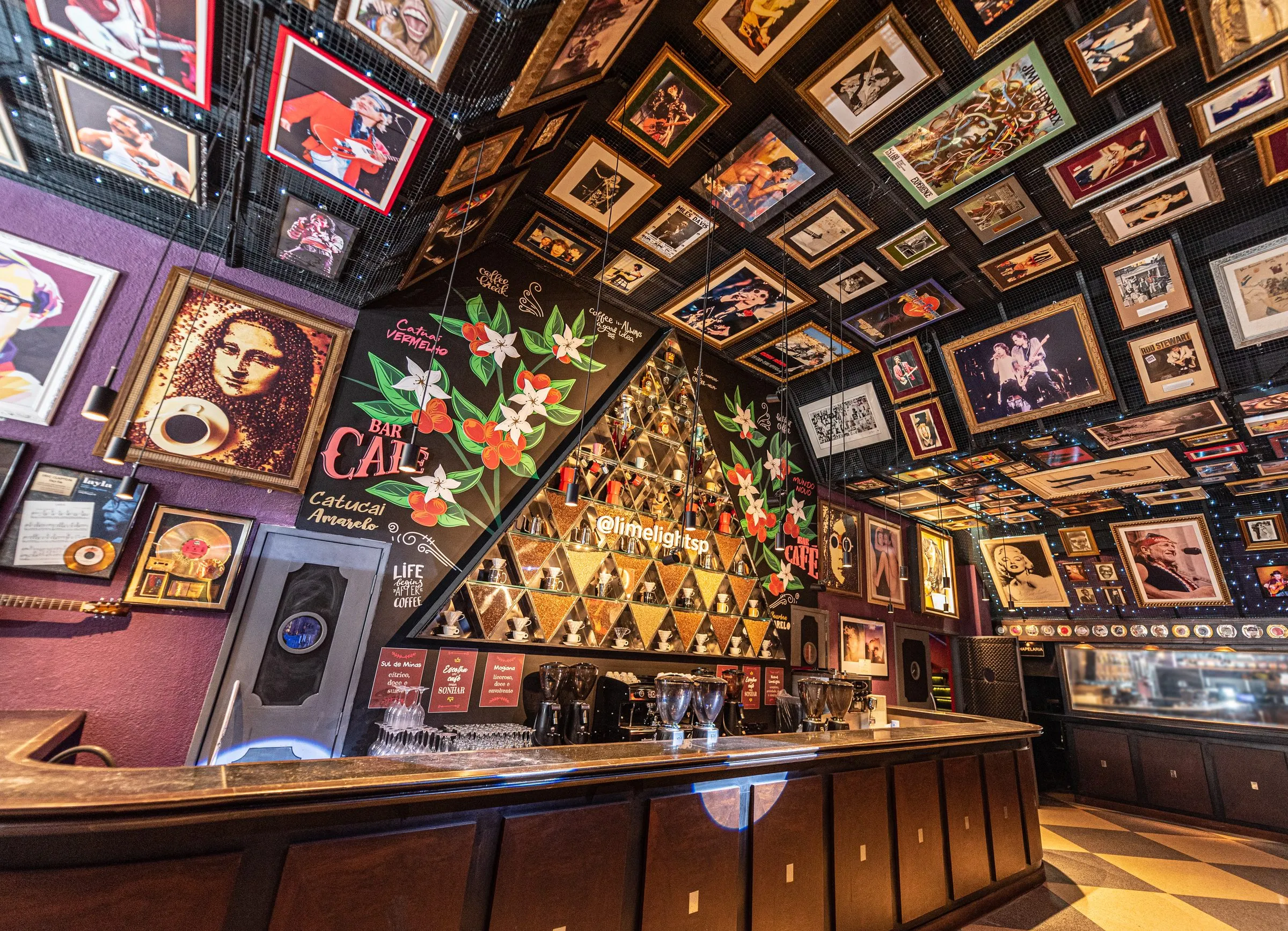Bar Café da Limelight visto de baixo, com o teto e as paredes forrados de quadros de músicos, o mural de flores e frutos de café, a estante triangular de grãos e xícaras com o letreiro @limelightsp e o balcão de madeira escura