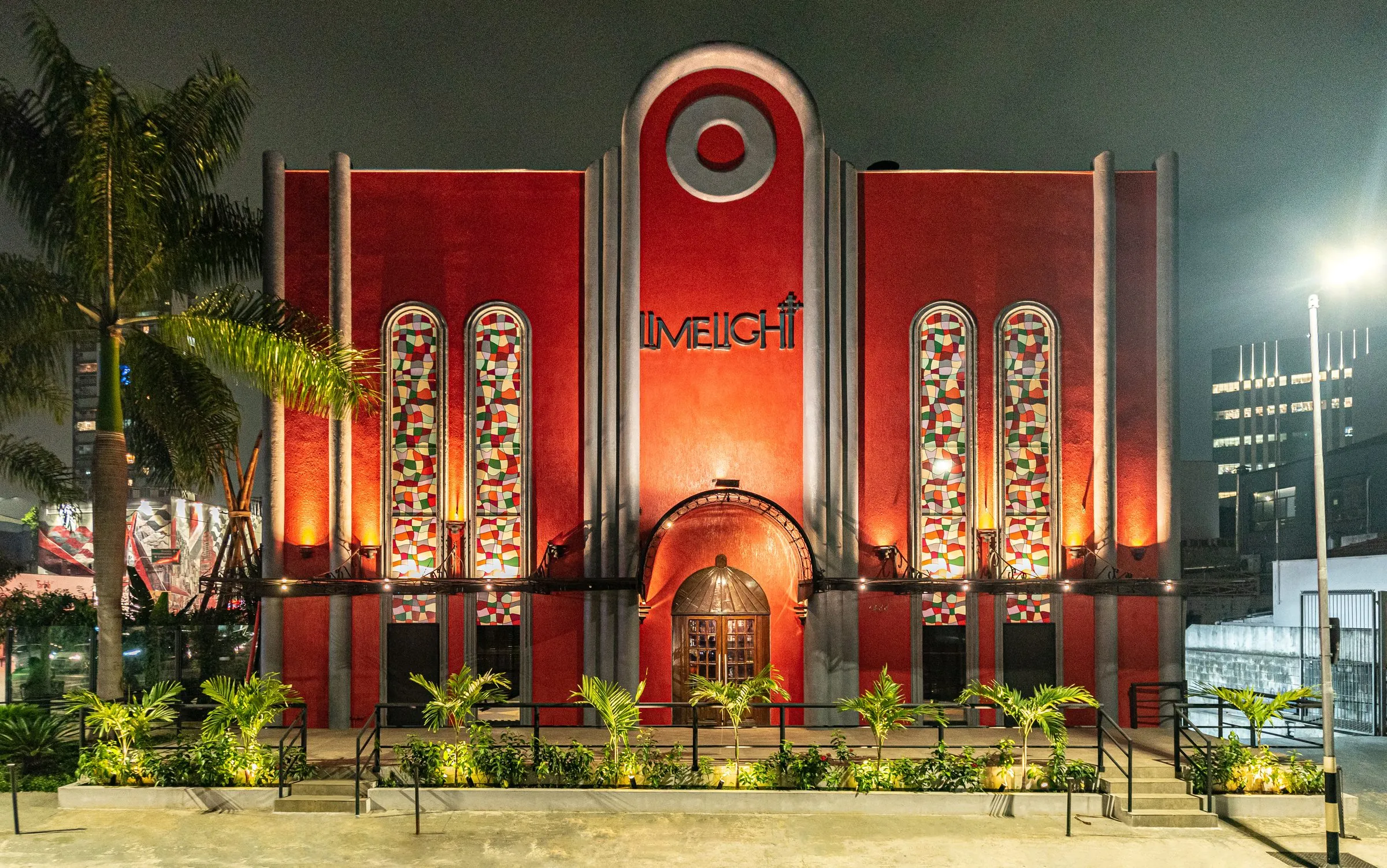 Fachada vermelha da Limelight à noite, na Vila Olímpia, em São Paulo, com quatro vitrais coloridos em arco, o letreiro Limelight no centro, a porta de madeira sob a marquise em arco e o canteiro de palmeiras iluminado na frente