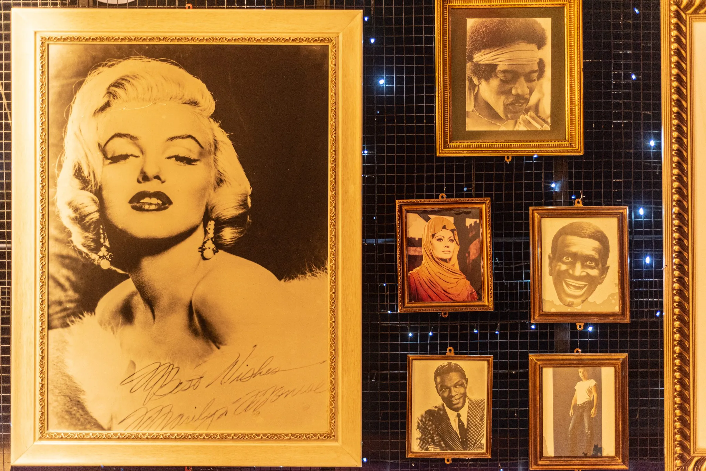 Foto autografada de Marilyn Monroe em moldura dourada ao lado de retratos de artistas em preto e branco, sobre uma parede escura com pontos de luz