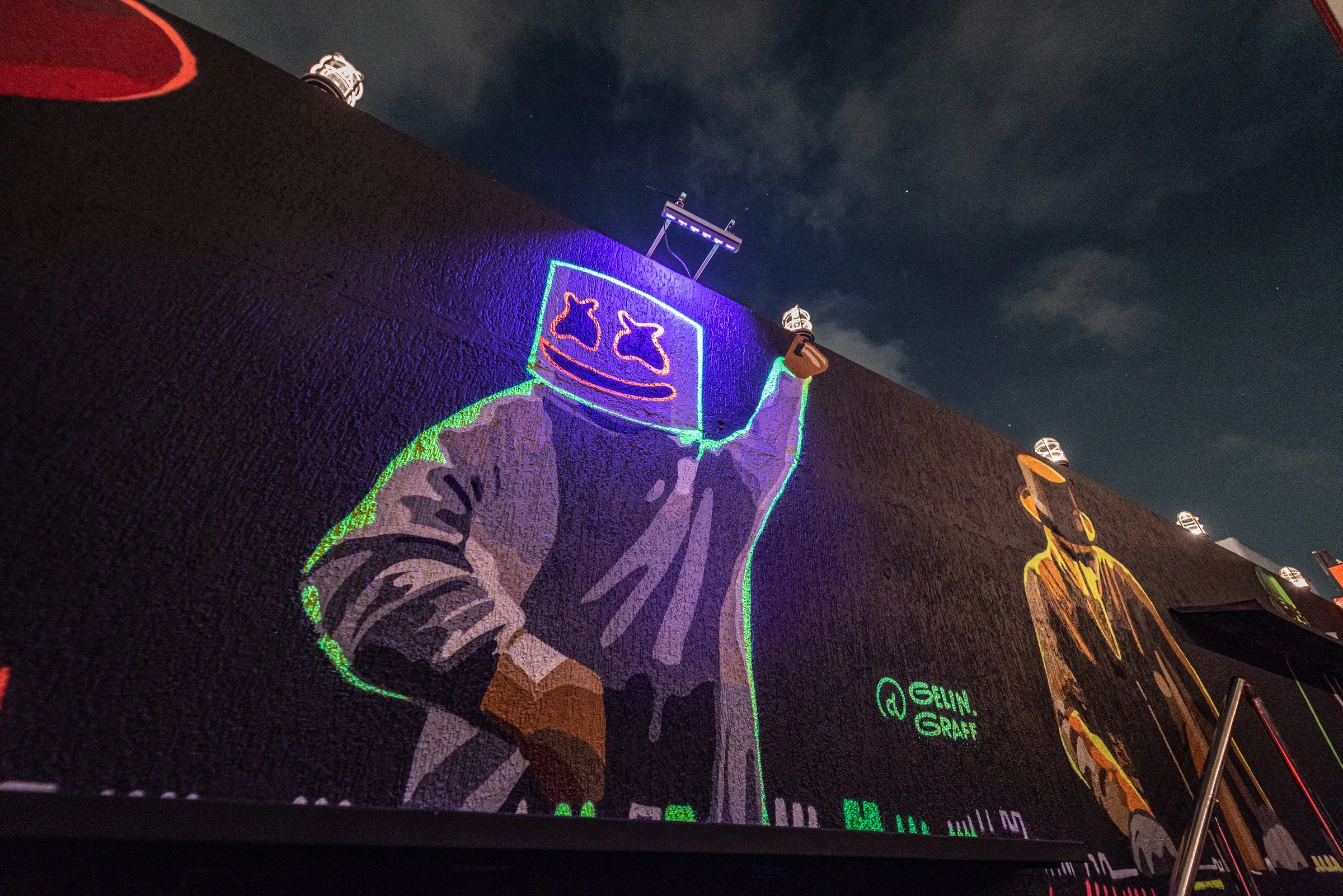 Mural grafitado na parede externa da Limelight à noite, com uma figura de capacete contornada em neon azul e verde e a assinatura do grafiteiro