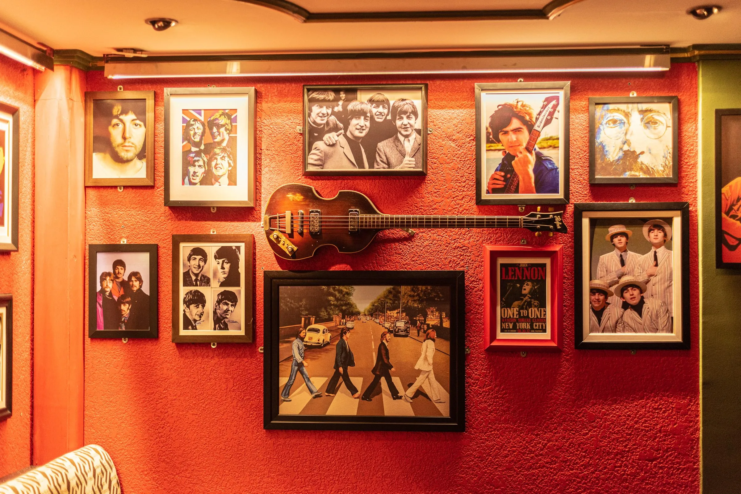 Parede vermelha com fotos emolduradas dos Beatles, entre elas a travessia da Abbey Road, um cartaz de Lennon e um baixo em forma de violino pendurado