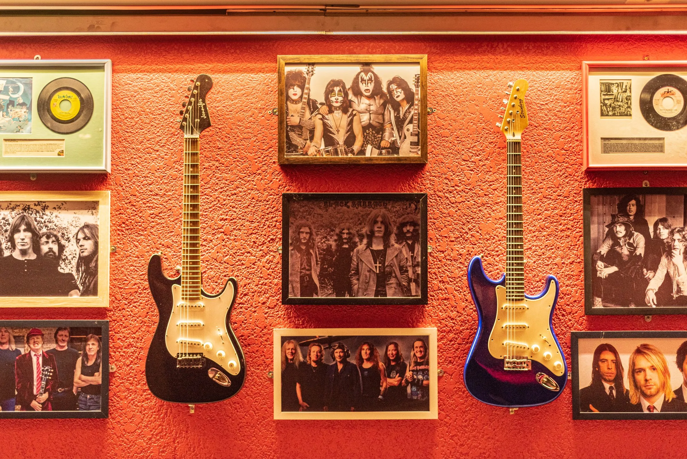Parede vermelha com duas guitarras, uma preta e uma azul, entre fotos em preto e branco de bandas de rock e discos emoldurados