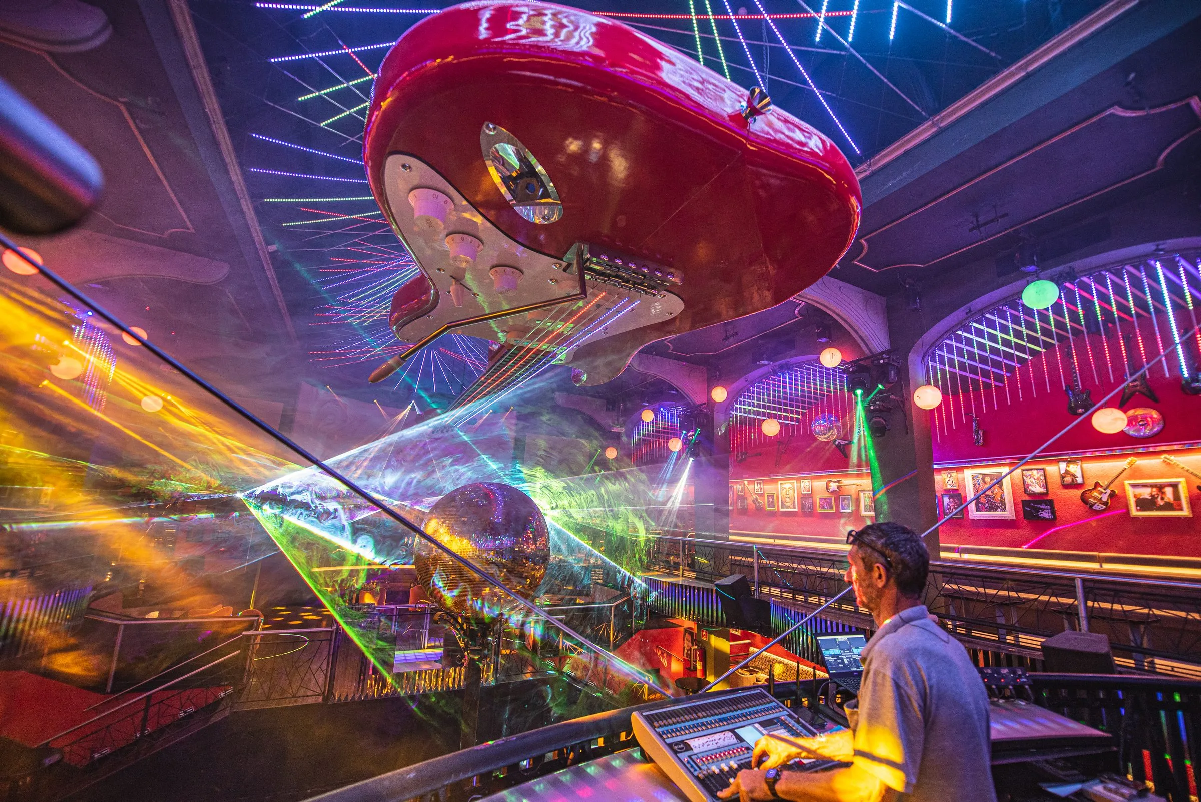 Guitarra vermelha gigante suspensa sobre a pista da Limelight, vista de baixo, com linhas de luz coloridas no teto, lasers verdes e amarelos atravessando a fumaça, o globo espelhado, as paredes vermelhas do mezanino com guitarras e quadros e um operador na mesa de luz em primeiro plano