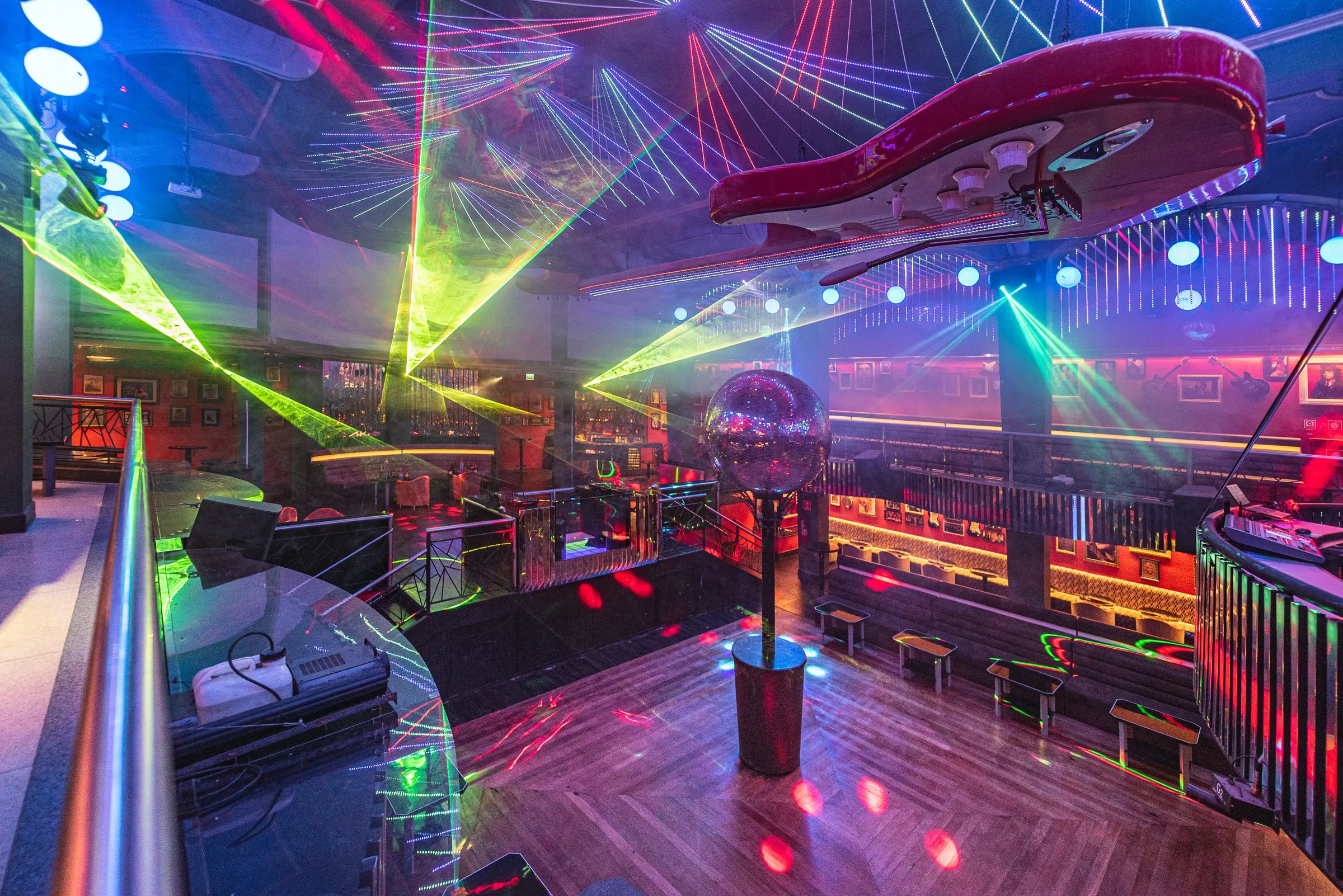 Lasers verdes e vermelhos cruzando a pista vista do mezanino, com o globo espelhado, a guitarra vermelha gigante no teto e os bancos ao redor do piso de madeira