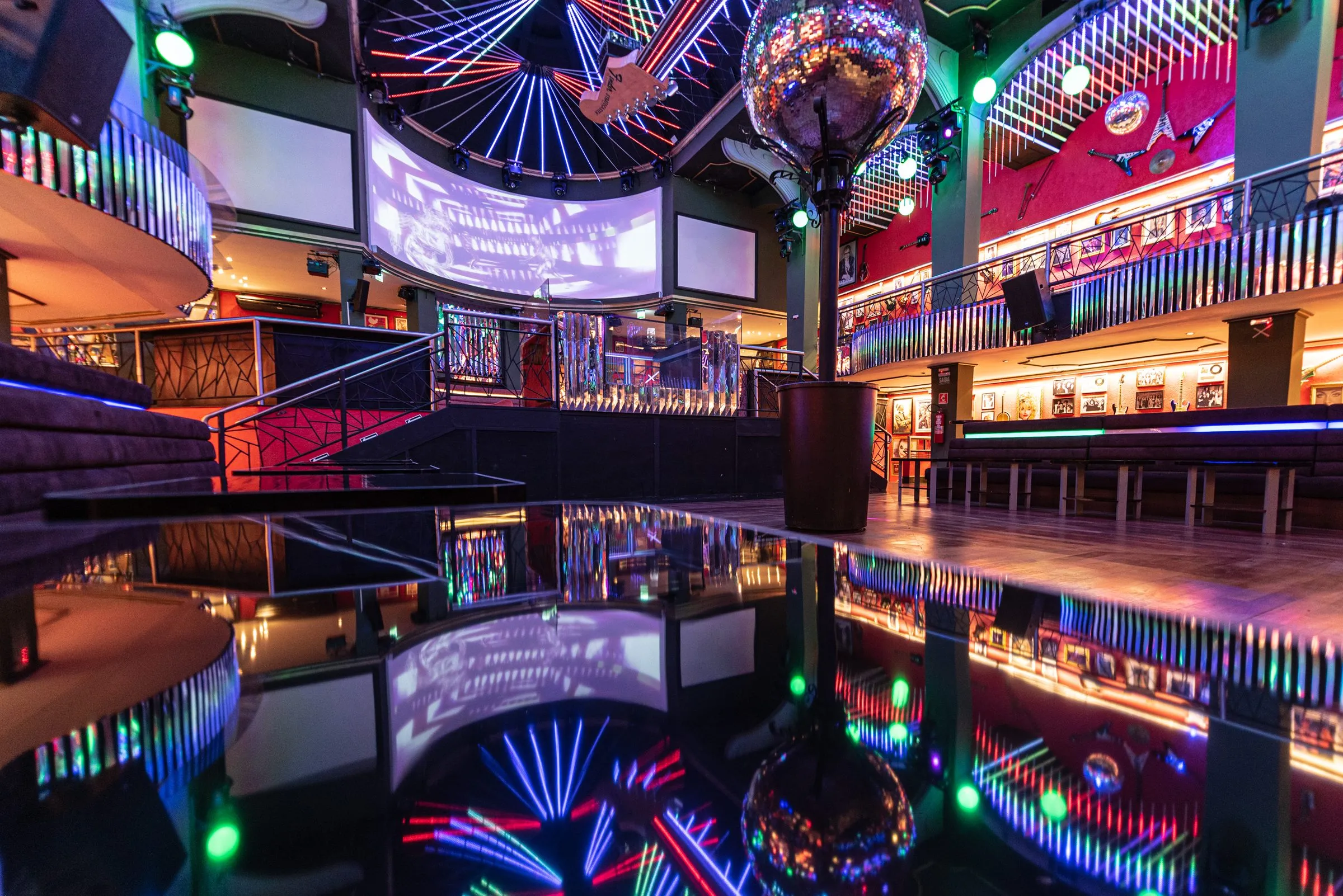 Pista da Limelight vista rente a uma mesa espelhada, que reflete o globo, o teto de luzes em leque, os telões curvos sobre o palco e os mezaninos iluminados