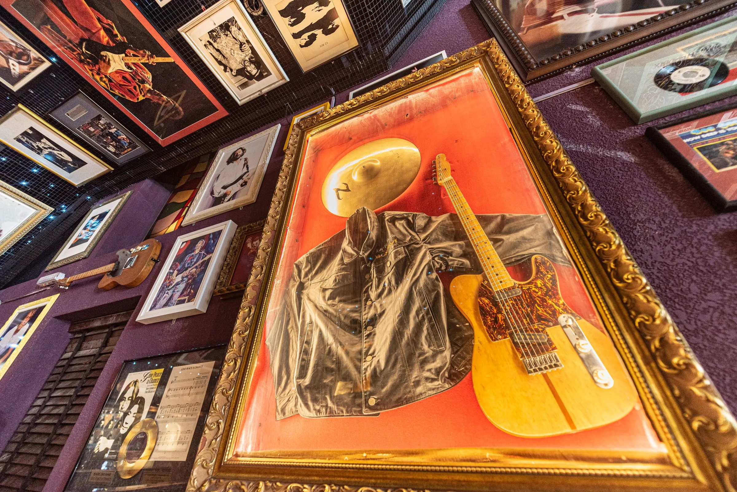Quadro de moldura dourada com uma jaqueta, uma guitarra de corpo claro e um prato de bateria sobre fundo laranja, numa parede roxa cheia de fotos e discos emoldurados