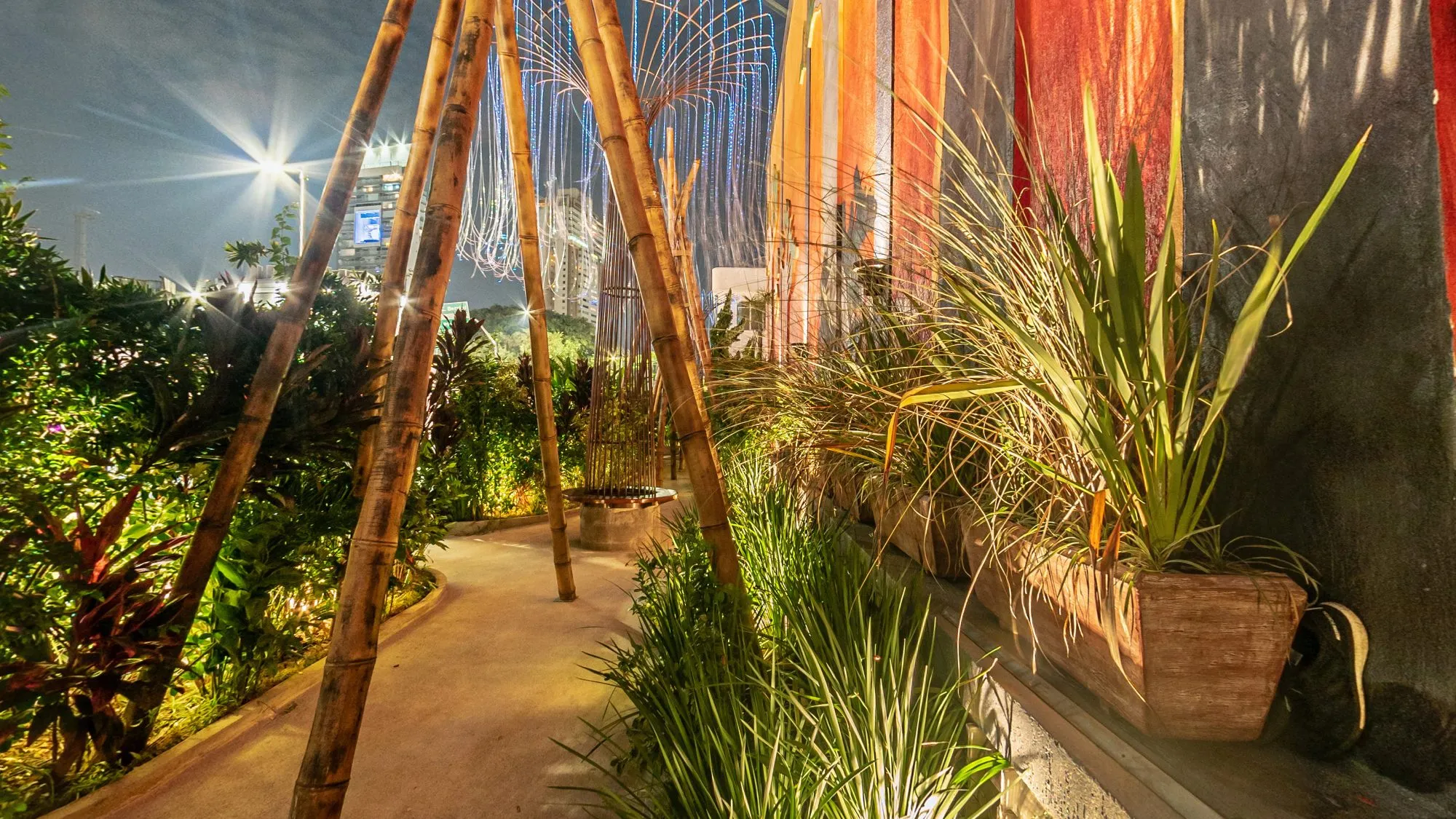 Jardim da Limelight à noite, com um caminho entre plantas iluminadas, estruturas de bambu, cortinas de luz e prédios da Vila Olímpia ao fundo