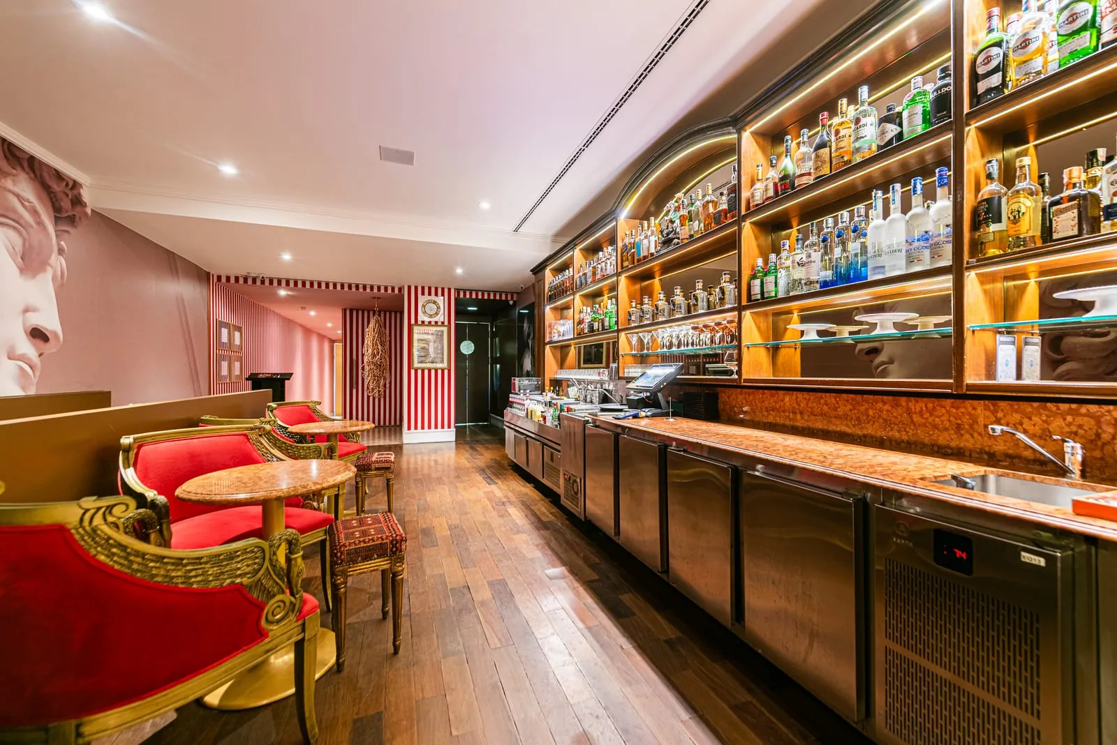 Bar com prateleiras de madeira e garrafas iluminadas, balcão de inox e poltronas douradas de veludo vermelho diante de um painel com rosto de estátua clássica