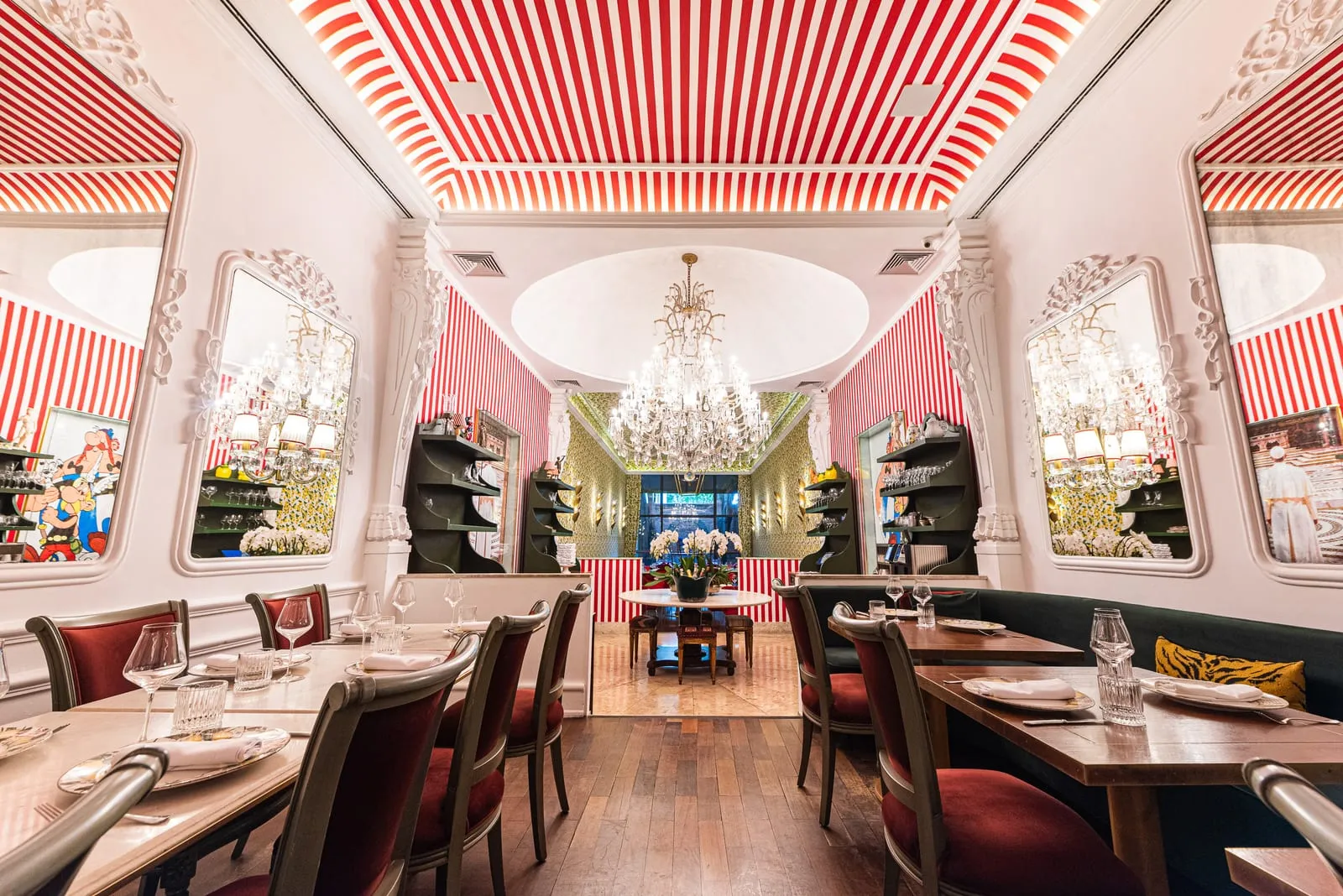 Mesas entre espelhos levando a uma sala com cúpula branca, lustre de cristal, paredes listradas e mesa redonda com orquídeas