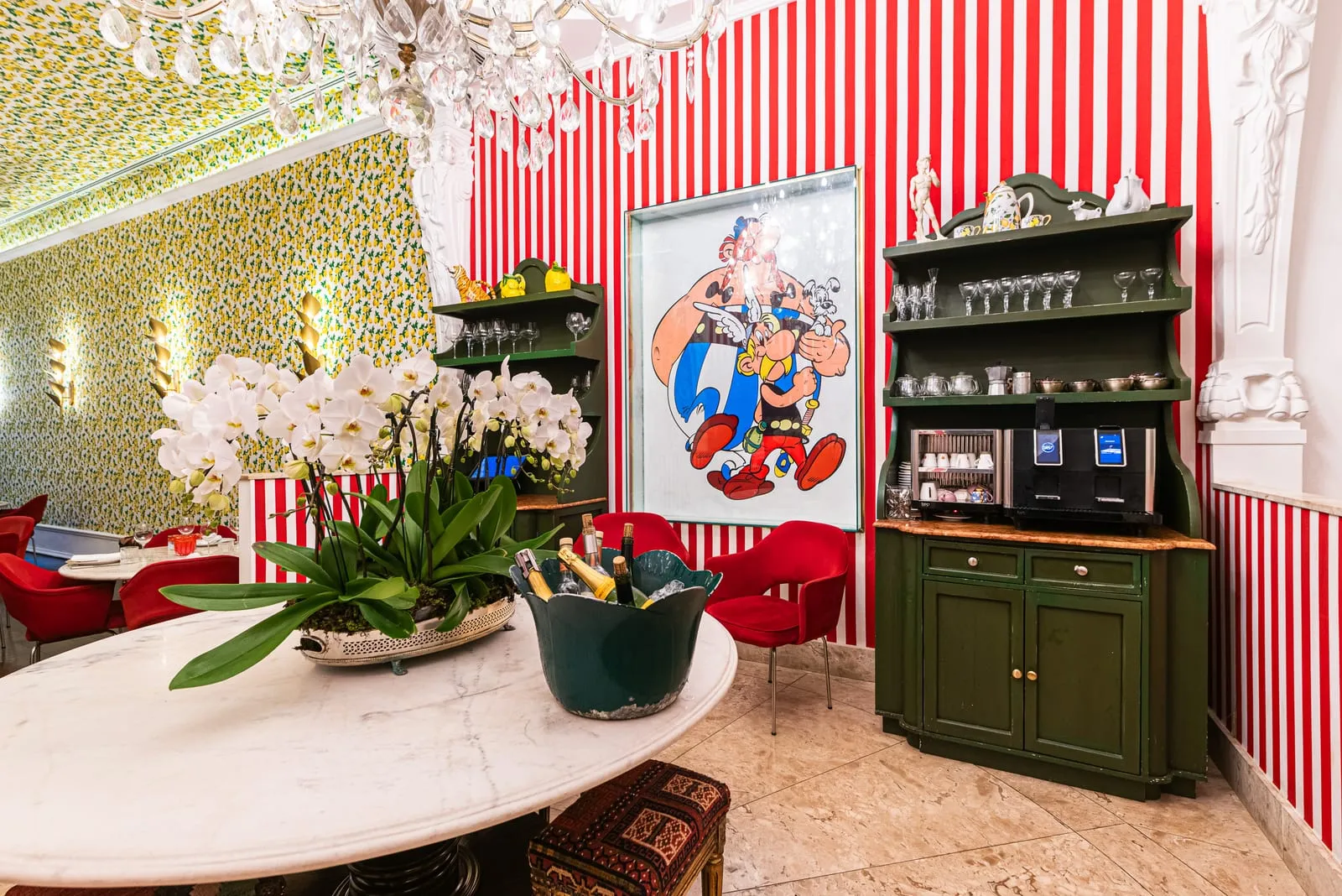 Mesa redonda de mármore com orquídeas brancas e balde de champanhe, aparador verde com louças e pôster do Asterix na parede listrada de vermelho