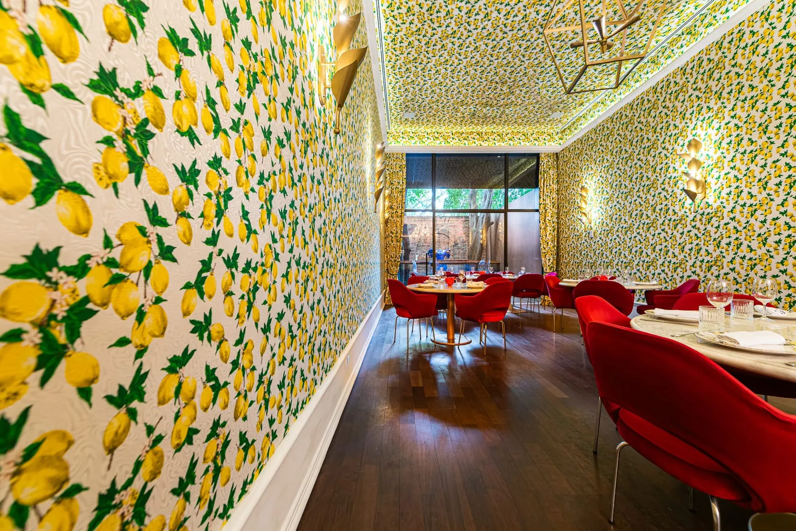 Papel de parede de limões amarelos e folhas verdes em primeiro plano, com mesas postas e cadeiras de veludo vermelho ao longo da sala