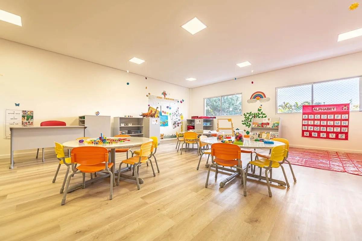 Sala de aula com mesas baixas, cadeiras laranja, armários e janelas amplas