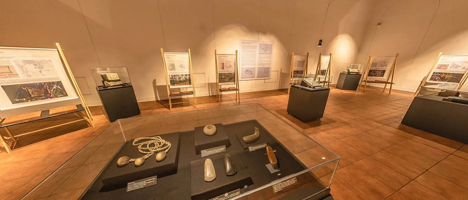 Vitrine com objetos de pedra e uma corda com pesos em primeiro plano, com painéis em cavaletes ao fundo da mostra Fortes Espanhóis