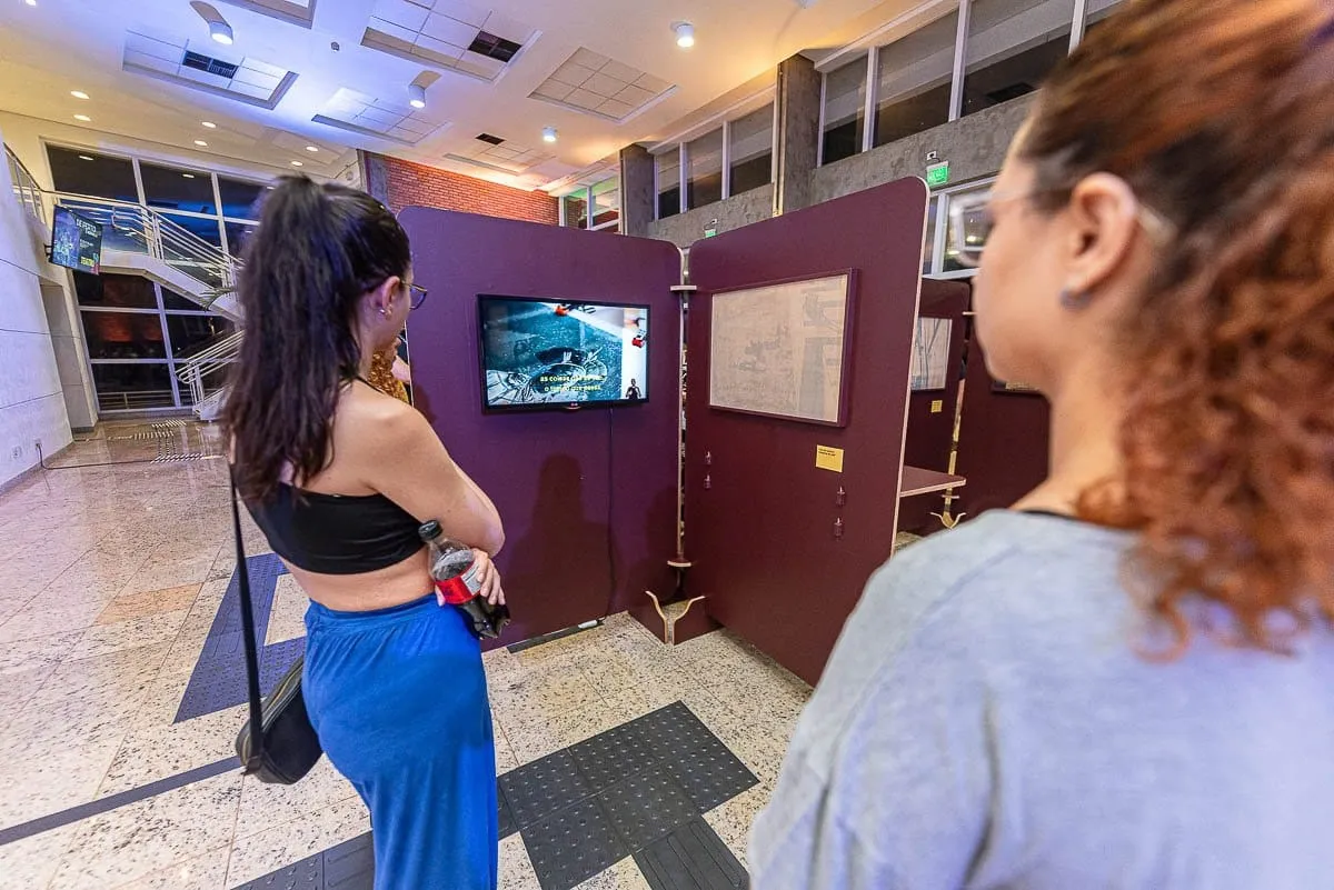 Duas visitantes assistem a um vídeo em tela instalada em um dos painéis da exposição
