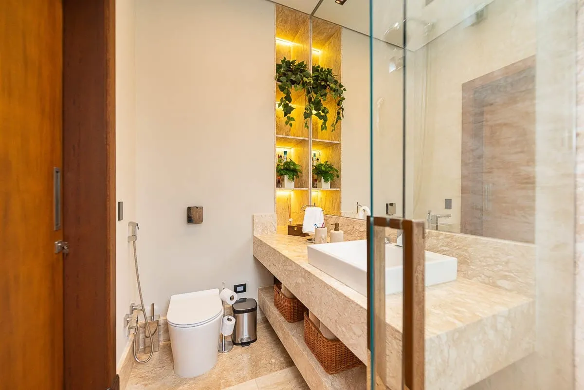 Lavabo com bancada esculpida em rocha clara, nichos iluminados com plantas e porta de vidro