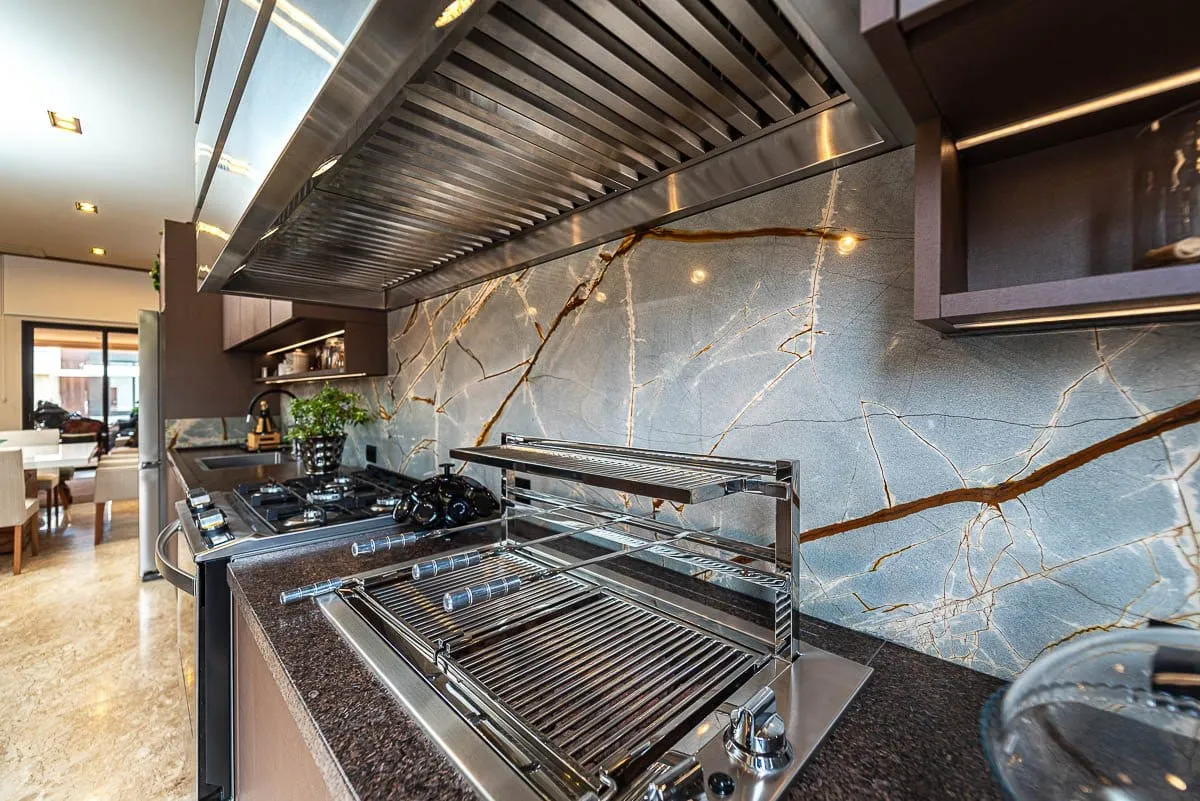 Cozinha gourmet com churrasqueira, cooktop e frontão em rocha verde, com a sala ao fundo
