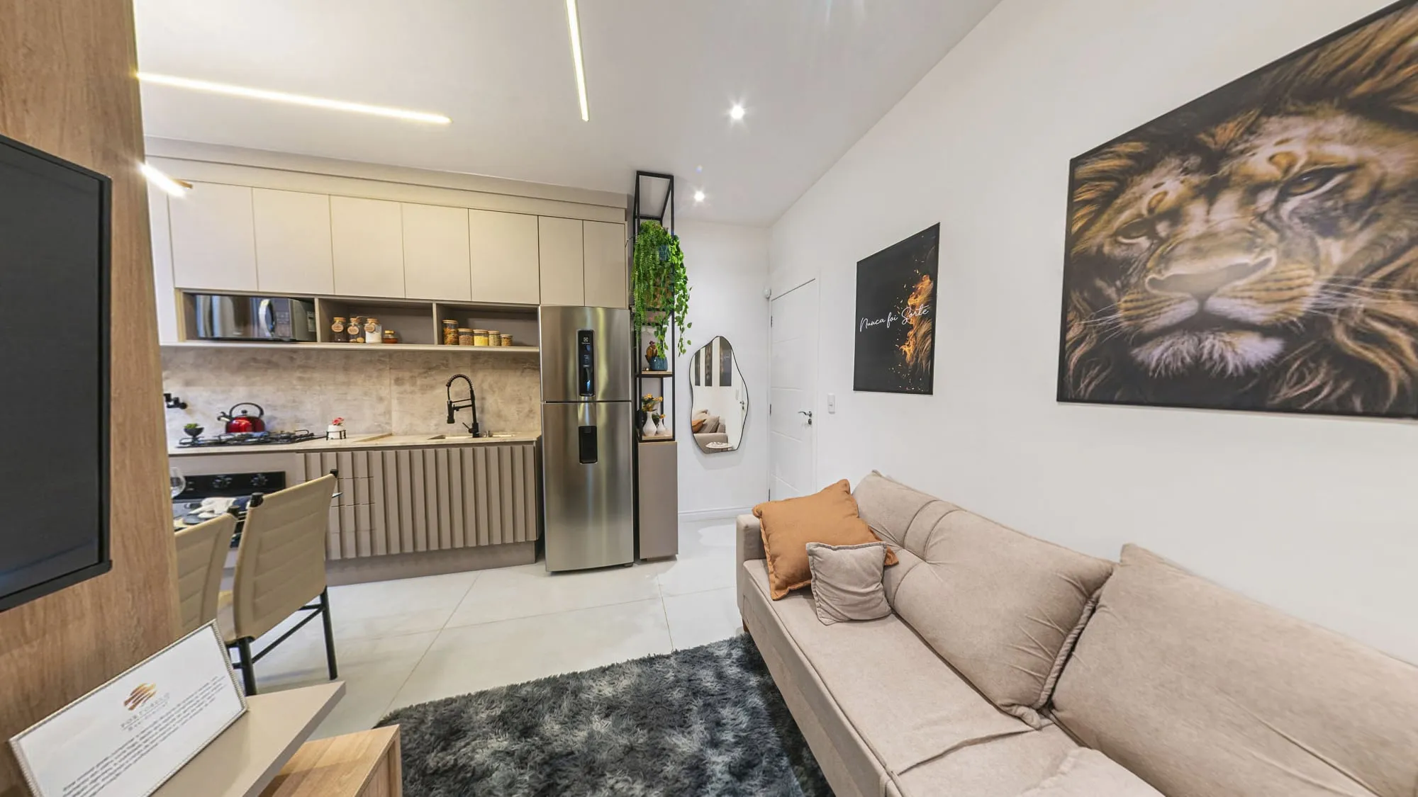 Sala de estar com sofá claro, tapete cinza felpudo e quadro de leão, com a cozinha planejada e a geladeira de inox ao fundo