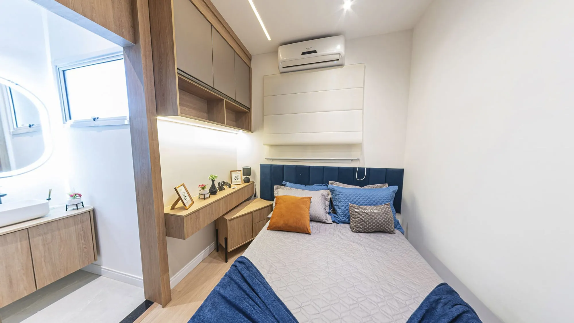 Suíte com cama de casal, cabeceira estofada azul, escrivaninha suspensa em madeira e a porta do banheiro da suíte à esquerda