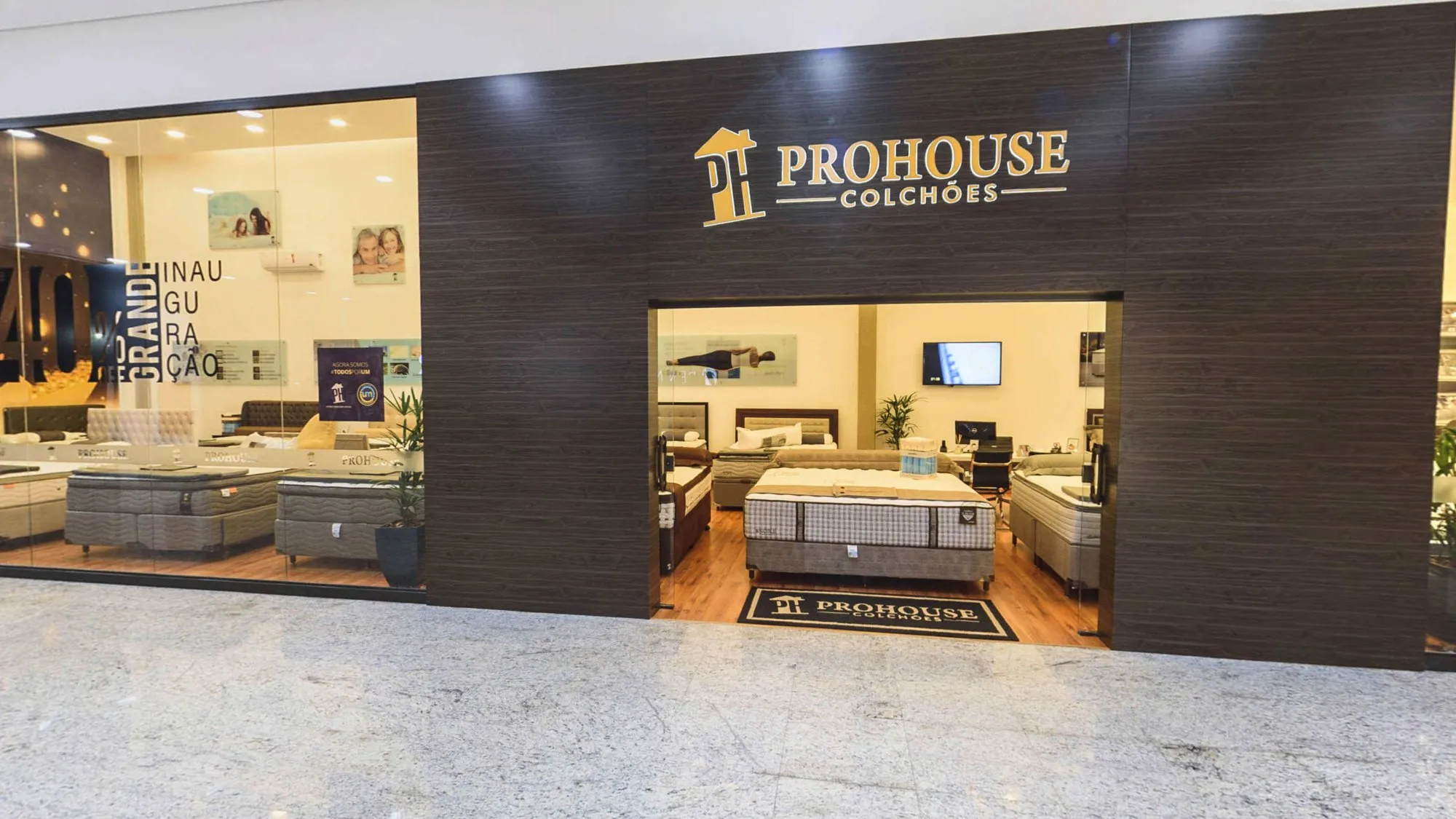 Fachada da Prohouse Colchões no corredor do Itapê Shopping, com revestimento de madeira escura, o logotipo dourado sobre a entrada, o tapete com a marca na porta e vitrines de vidro com camas box expostas