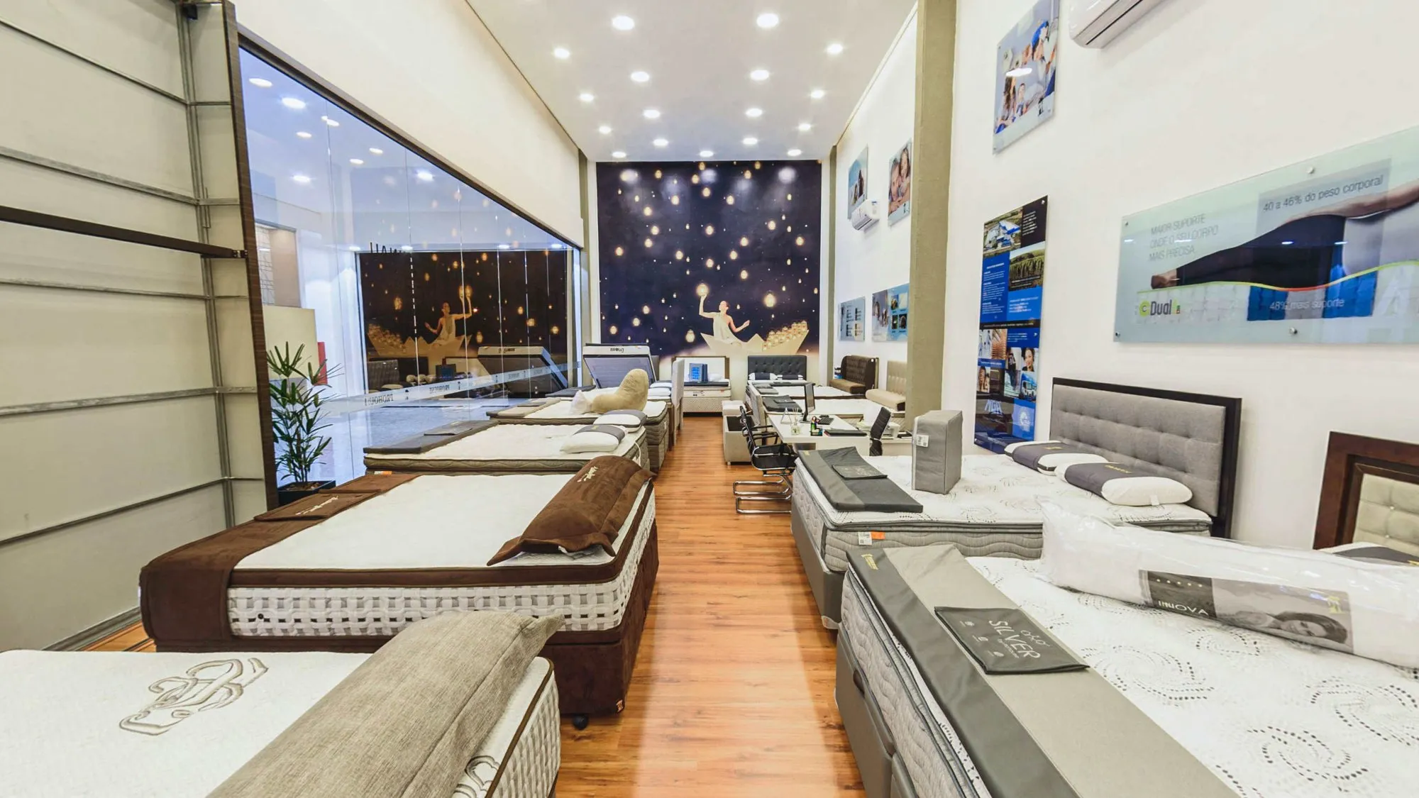 Salão da loja do Itapê Shopping visto da frente, com camas box de vários modelos dos dois lados do corredor de piso de madeira, a vitrine para o shopping à esquerda e o grande painel de céu estrelado na parede do fundo