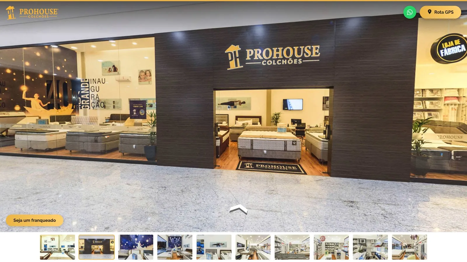 Cena do corredor do shopping no tour, com a fachada da Prohouse em madeira escura, o logotipo dourado sobre a entrada, as vitrines com camas expostas, a seta de navegação no piso e a miniatura da fachada destacada na galeria