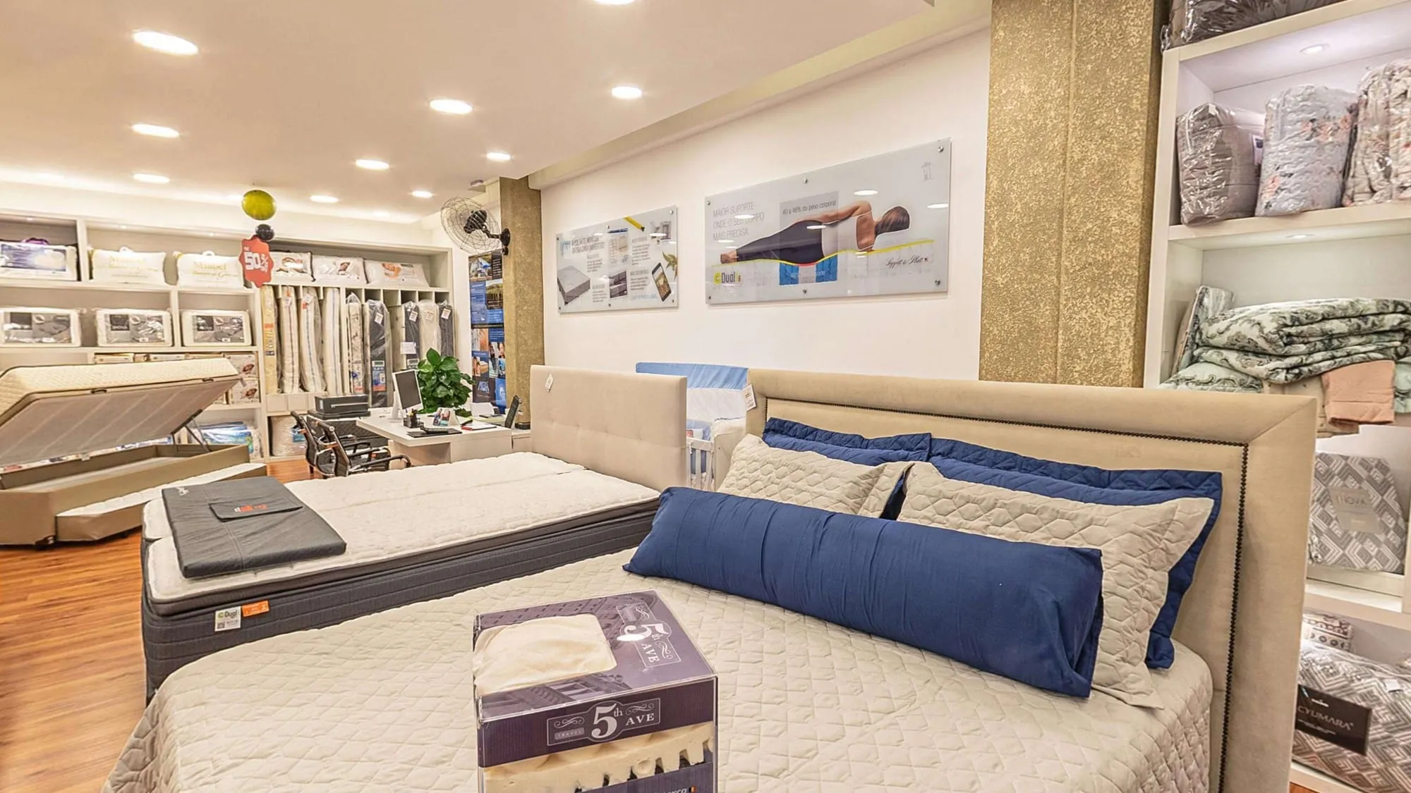 Cama montada com cabeceira estofada bege, colcha clara e almofadas azul-marinho, com nichos de travesseiros e roupa de cama e quadros de produtos na parede ao fundo