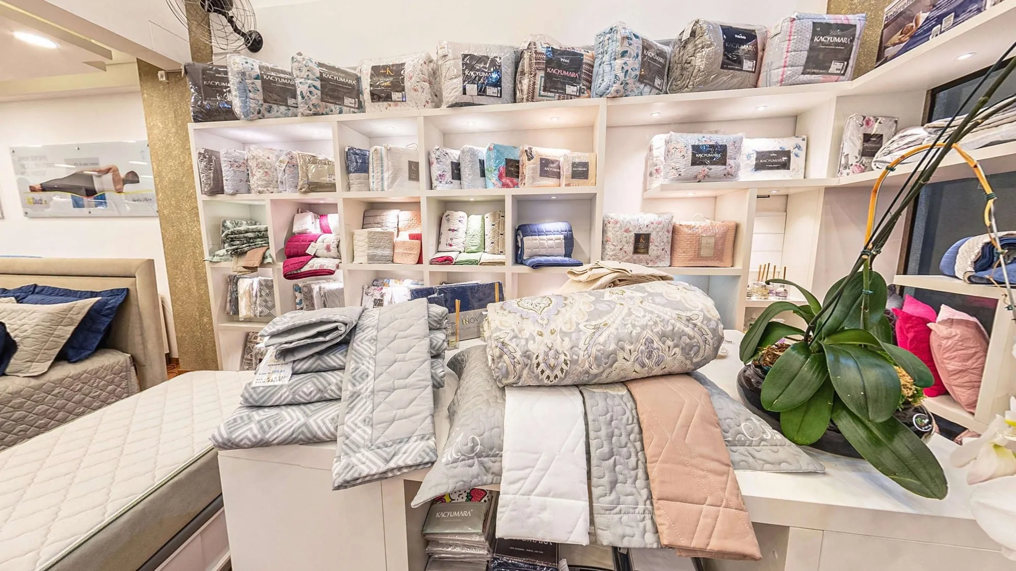 Ilha de roupa de cama com edredons e colchas dobrados e uma orquídea, diante de estantes brancas cheias de jogos de cama, travesseiros e almofadas coloridas