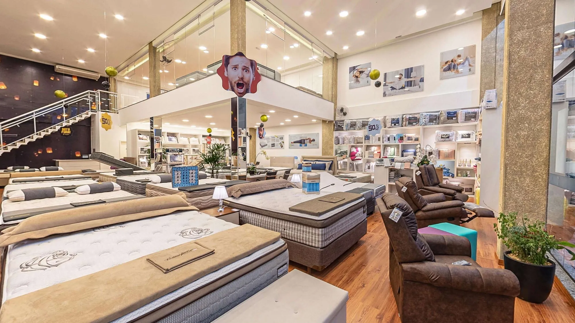 Salão da matriz da Prohouse Colchões, com fileiras de colchões e camas box, poltronas reclináveis marrons à direita, estantes de roupa de cama ao fundo, o mezanino envidraçado e a escada branca diante da parede escura com lanternas