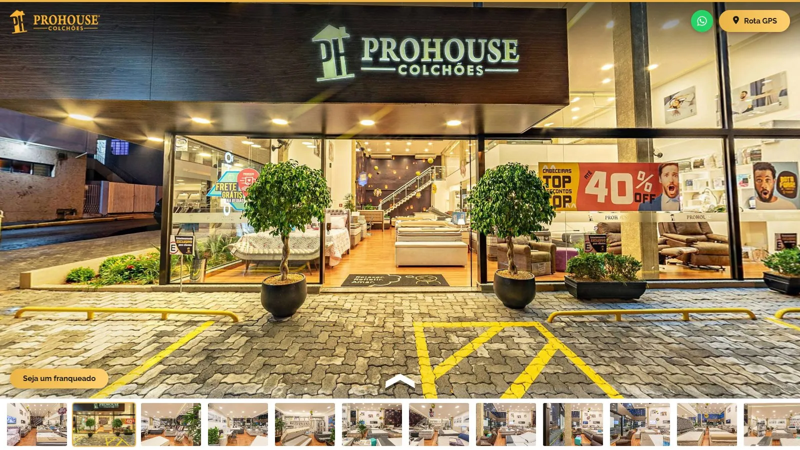 Cena externa do tour com a fachada da matriz da Prohouse Colchões à noite, o letreiro iluminado, a vitrine de vidro mostrando o salão, dois vasos com árvores na entrada, o estacionamento de piso intertravado e a seta de navegação para entrar na loja