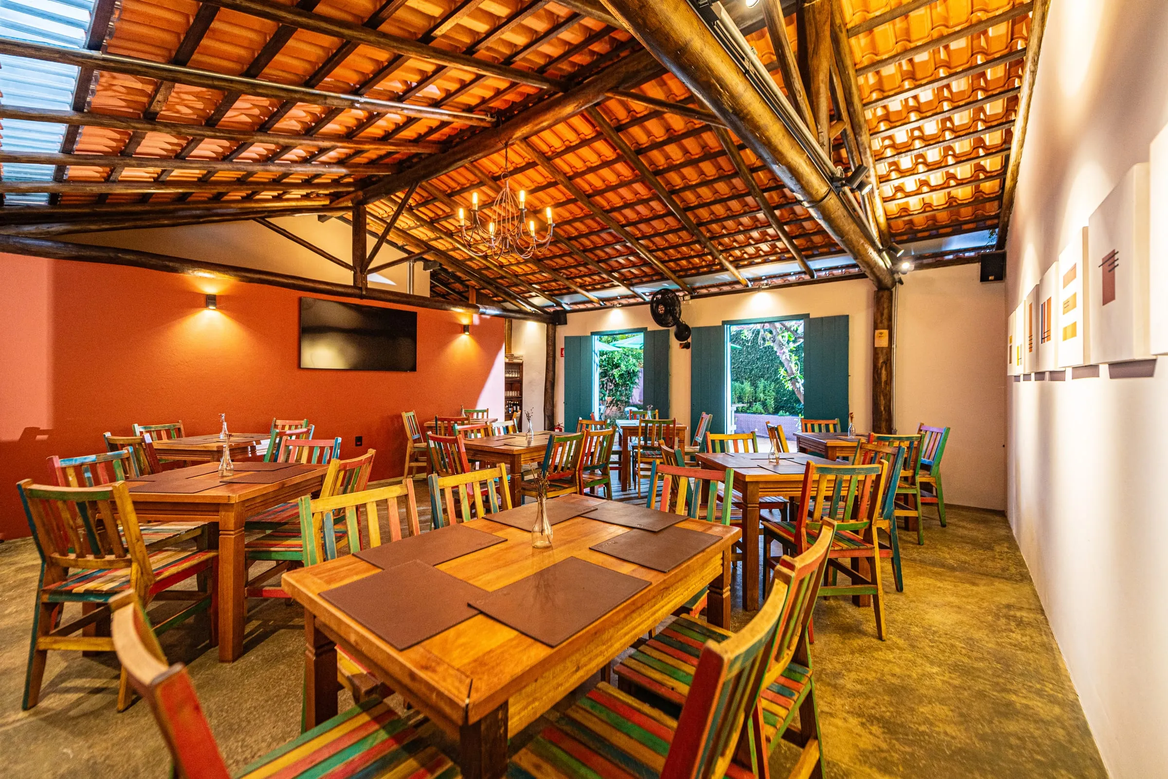 Salão Galeria do Quintal Iwasa'i, comprido, com parede laranja, lustre de ferro sob o telhado de telhas aparentes, mesas de madeira com jogos americanos de couro, cadeiras de ripas coloridas, quadros na parede branca à direita e duas portas azuis abertas para o jardim ao fundo