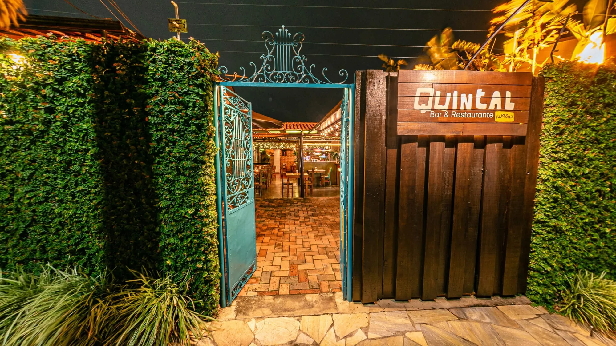 Fachada do Quintal Iwasa'i à noite no tour 360°, com o portão de ferro azul aberto para um corredor de tijolos que leva ao salão iluminado, a placa de madeira Quintal Bar & Restaurante e muros cobertos de hera