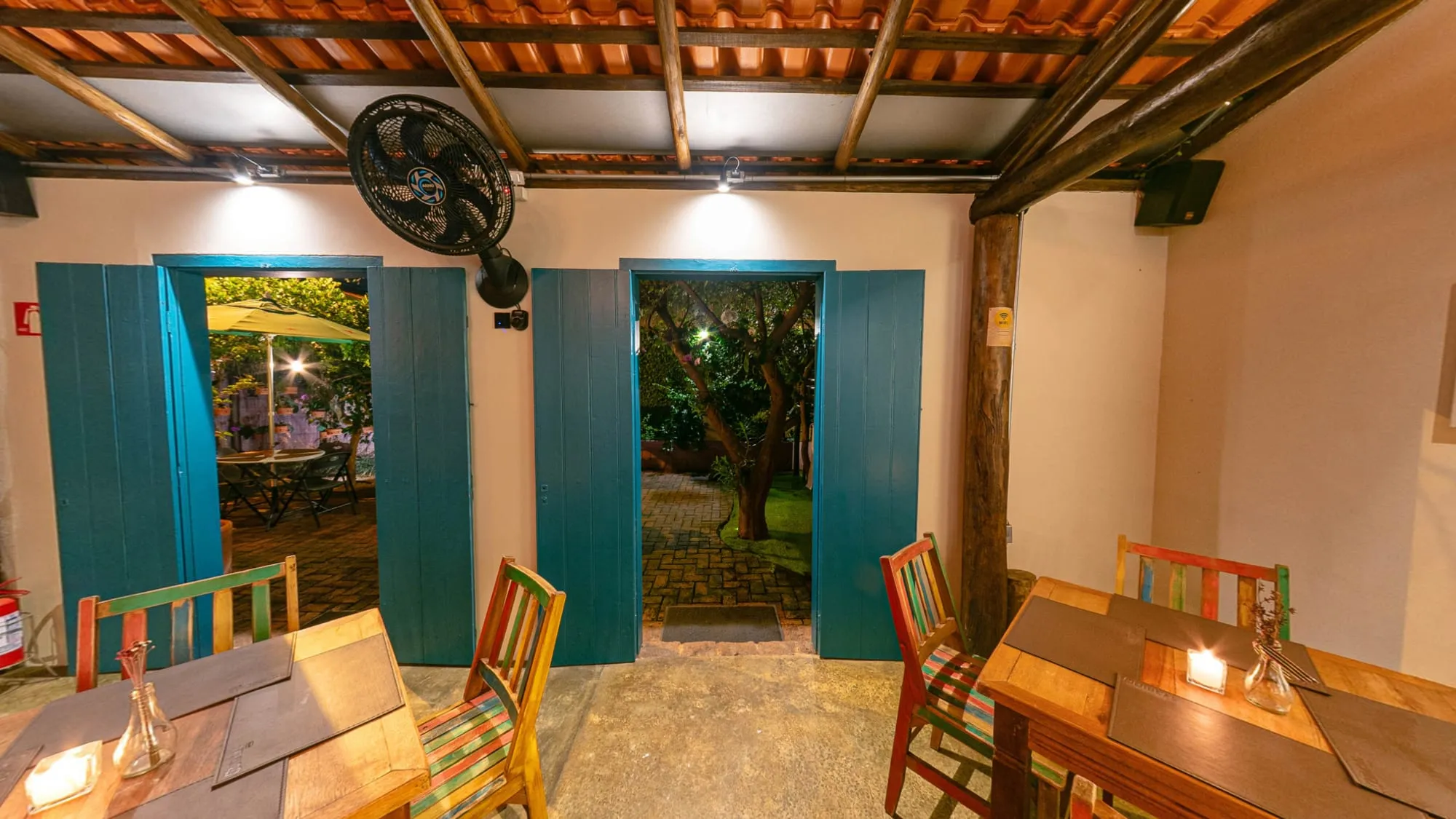Salão Galeria do Quintal Iwasa'i visto de dentro, com duas portas de madeira azul abertas para o jardim iluminado, guarda-sol e árvore lá fora, paredes claras, telhado de telha aparente e mesas de madeira com cadeiras de ripas coloridas e velas
