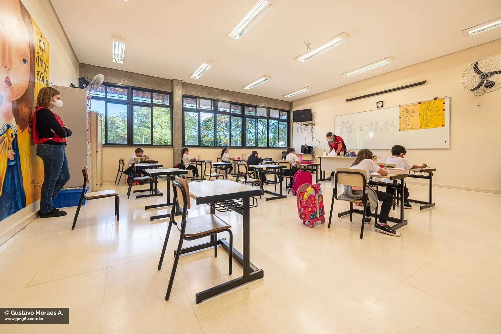 Sala de aula com alunos em carteiras espaçadas e professora ao fundo