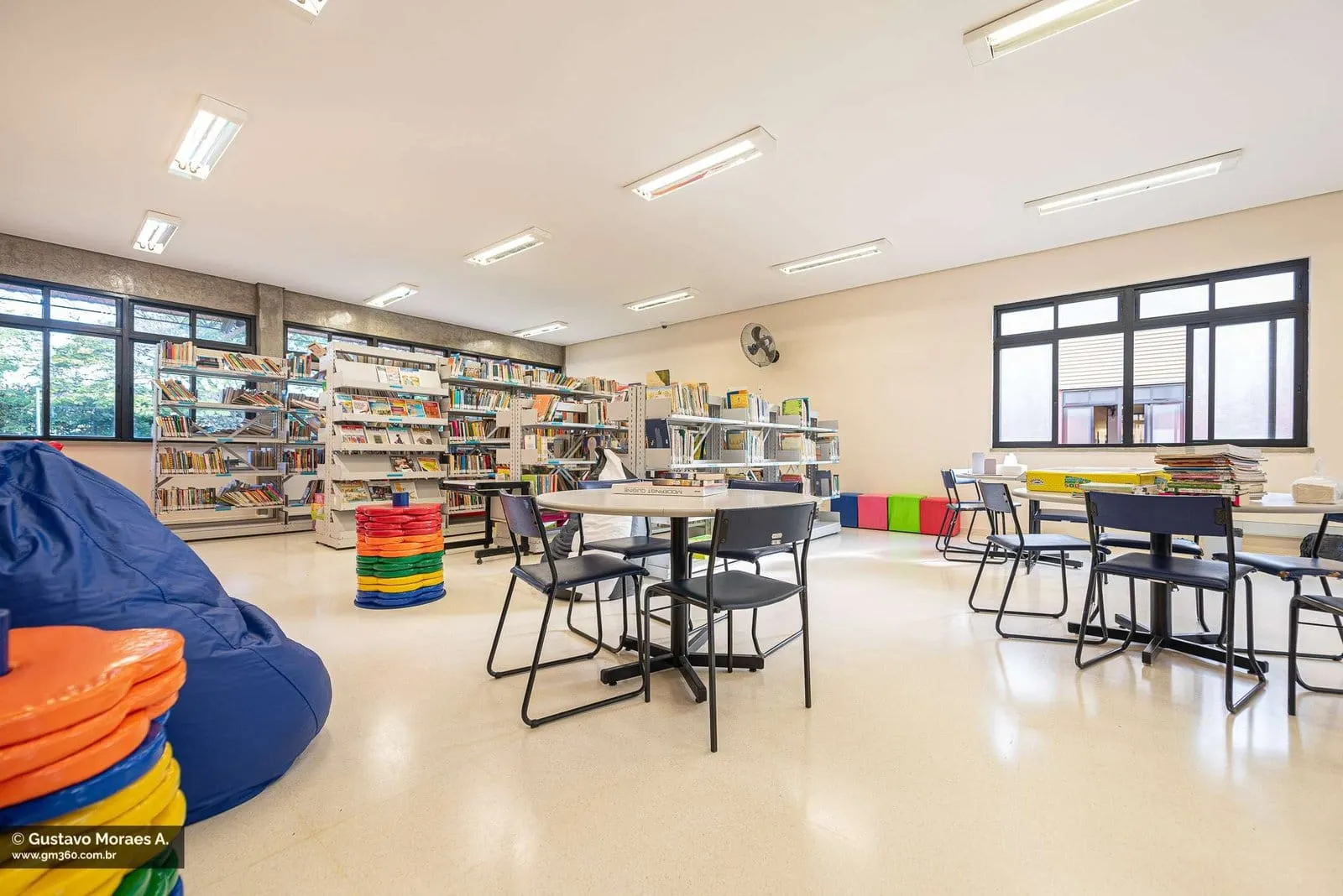 Biblioteca com mesas redondas, estantes de livros e pufes coloridos