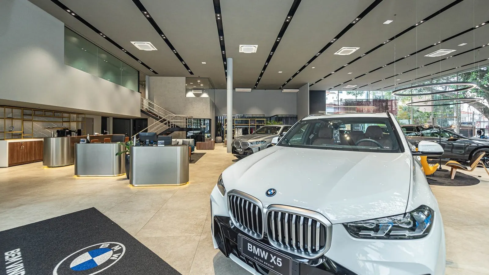 Salão da BMW Osten Perdizes com um BMW X5 branco em primeiro plano, balcões de atendimento e escada para o mezanino ao fundo